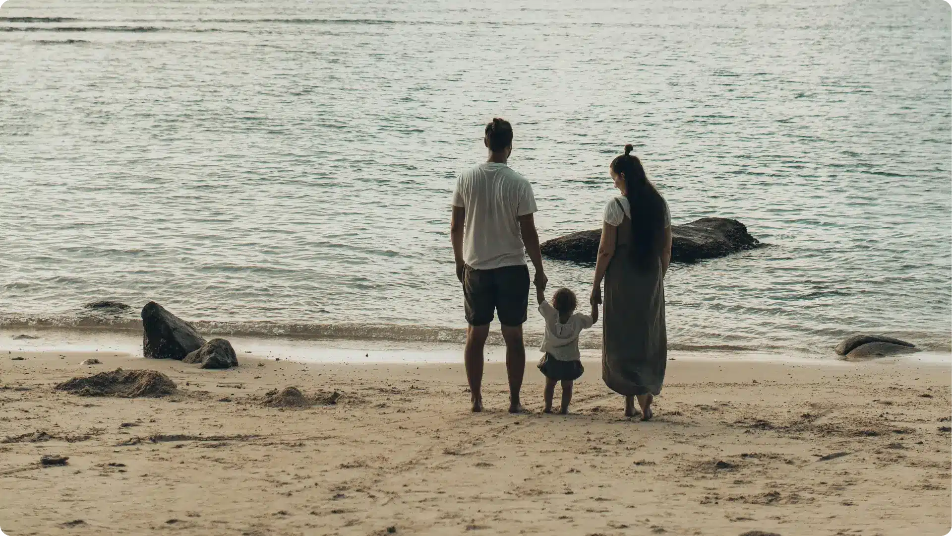 A mother and father holding their young child's hands on a beach