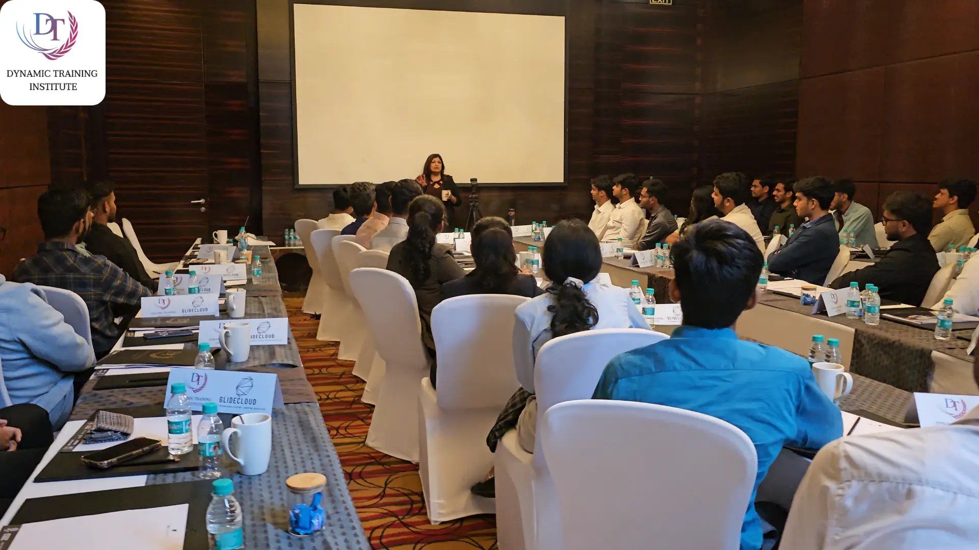 A woman trainer speaking at the front of a hotel conference room to participants seated along long tables with “Glide Cloud” name cards
