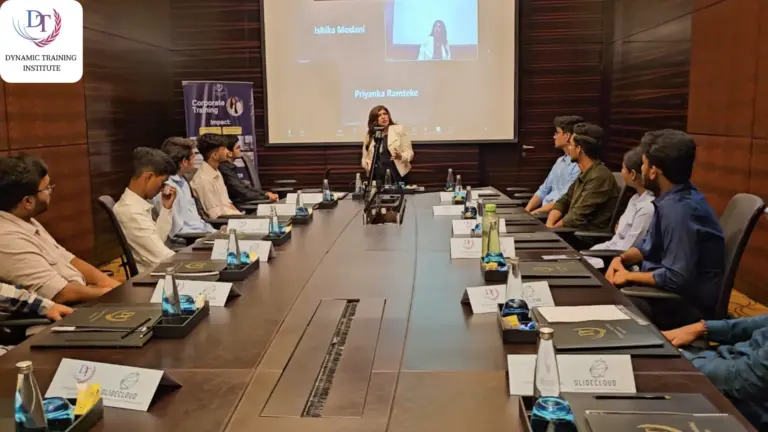 A woman trainer speaking at the head of a long boardroom table to participants seated on both sides