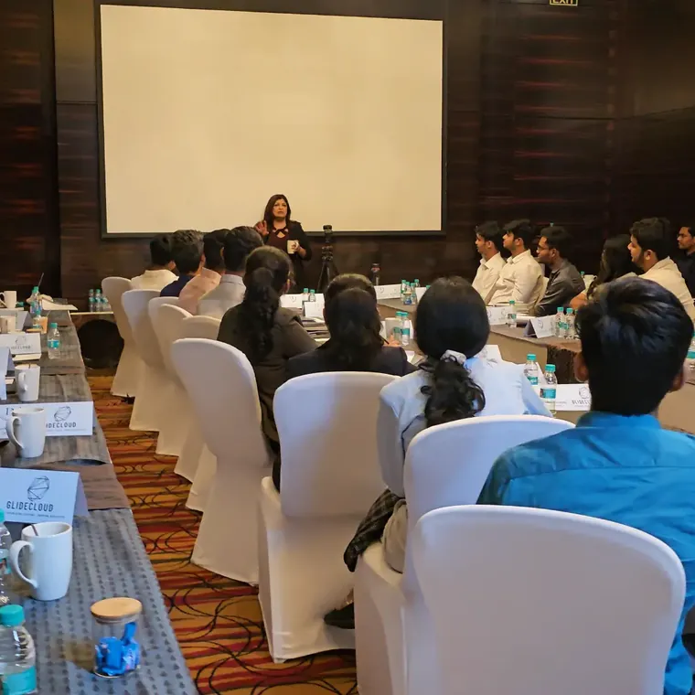 A woman trainer speaking at the front of a hotel conference room to participants seated along long tables with “Glide Cloud” name cards
