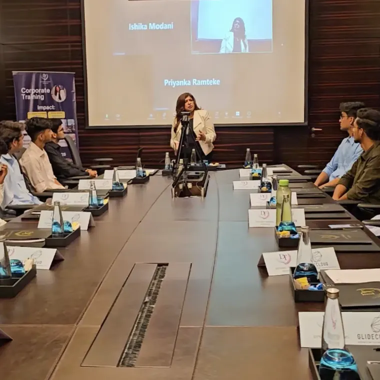 A woman trainer speaking at the head of a long boardroom table to participants seated on both sides