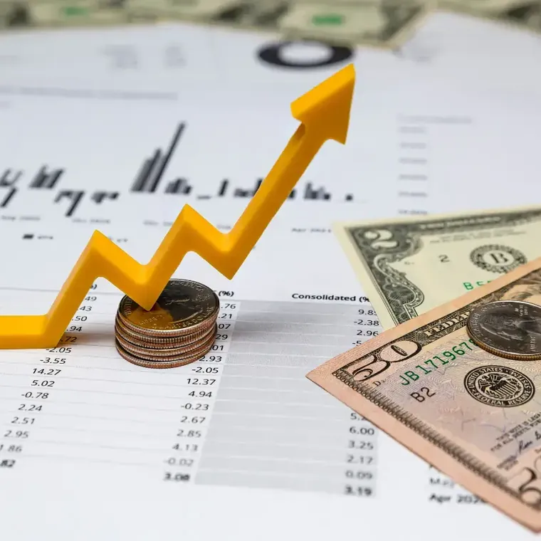 A rising yellow arrow drawn over a printed financial chart, with stacked coins and dollar notes