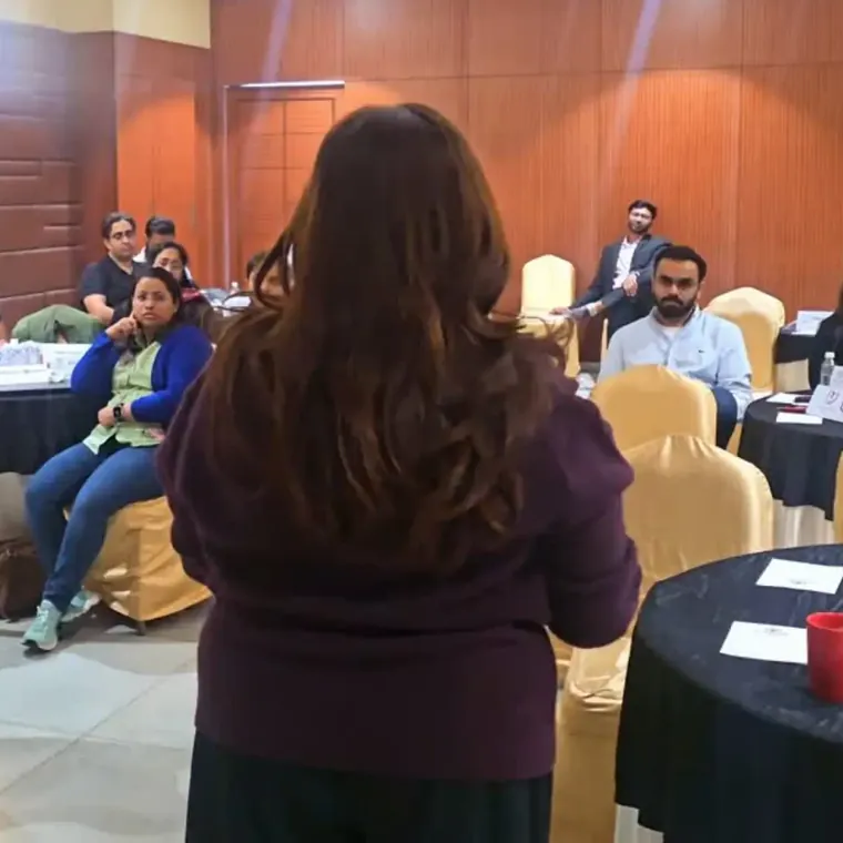 A trainer, seen from behind, speaking to participants seated at round tables in a hotel training room