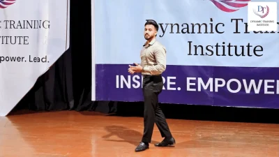 A young man in a white shirt speaking on stage in front of the Dynamic Training Institute banner
