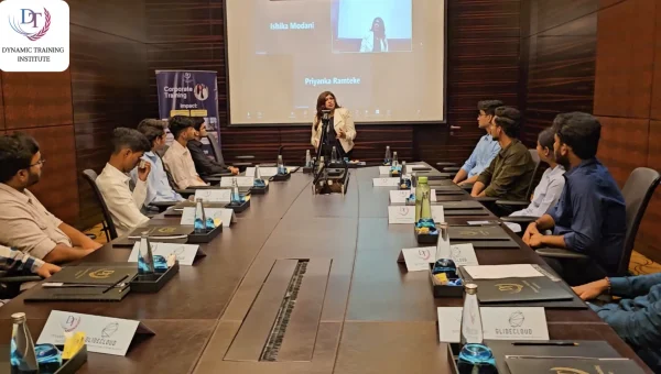 A woman trainer speaking at the head of a long boardroom table to participants seated on both sides