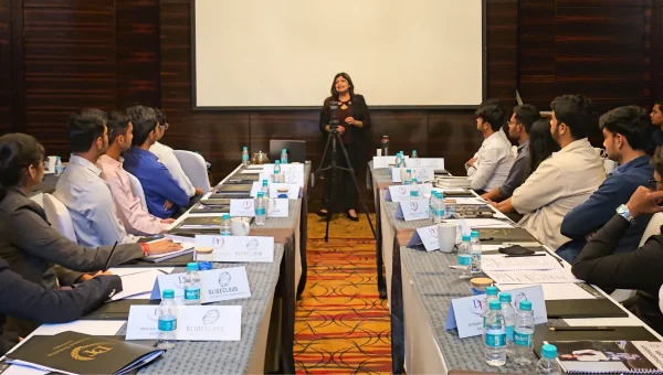 Tina Shah presenting at the head of two long tables to participants seated with name cards in a hotel conference room
