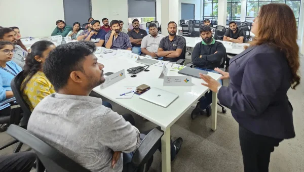A trainer in a dark blazer speaking to professionals seated around white tables in an office training room
