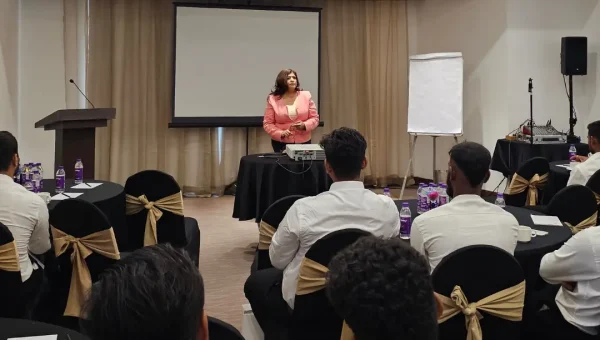 A woman trainer speaking from the front of a hotel conference room to participants seated at round tables